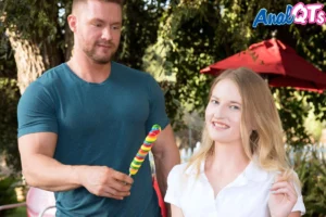 Smiling petite blonde Lolli Lane in white top holds giant rainbow lollipop outdoors next to muscular Sam Shock in teal shirt on sunny patio