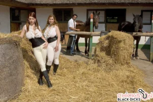 Two busty women in tight riding outfits pose in a hay-filled stable with horses; Christy Marks (left) and Terry Nova (right) stand in front of Carlos Rios handling the horses