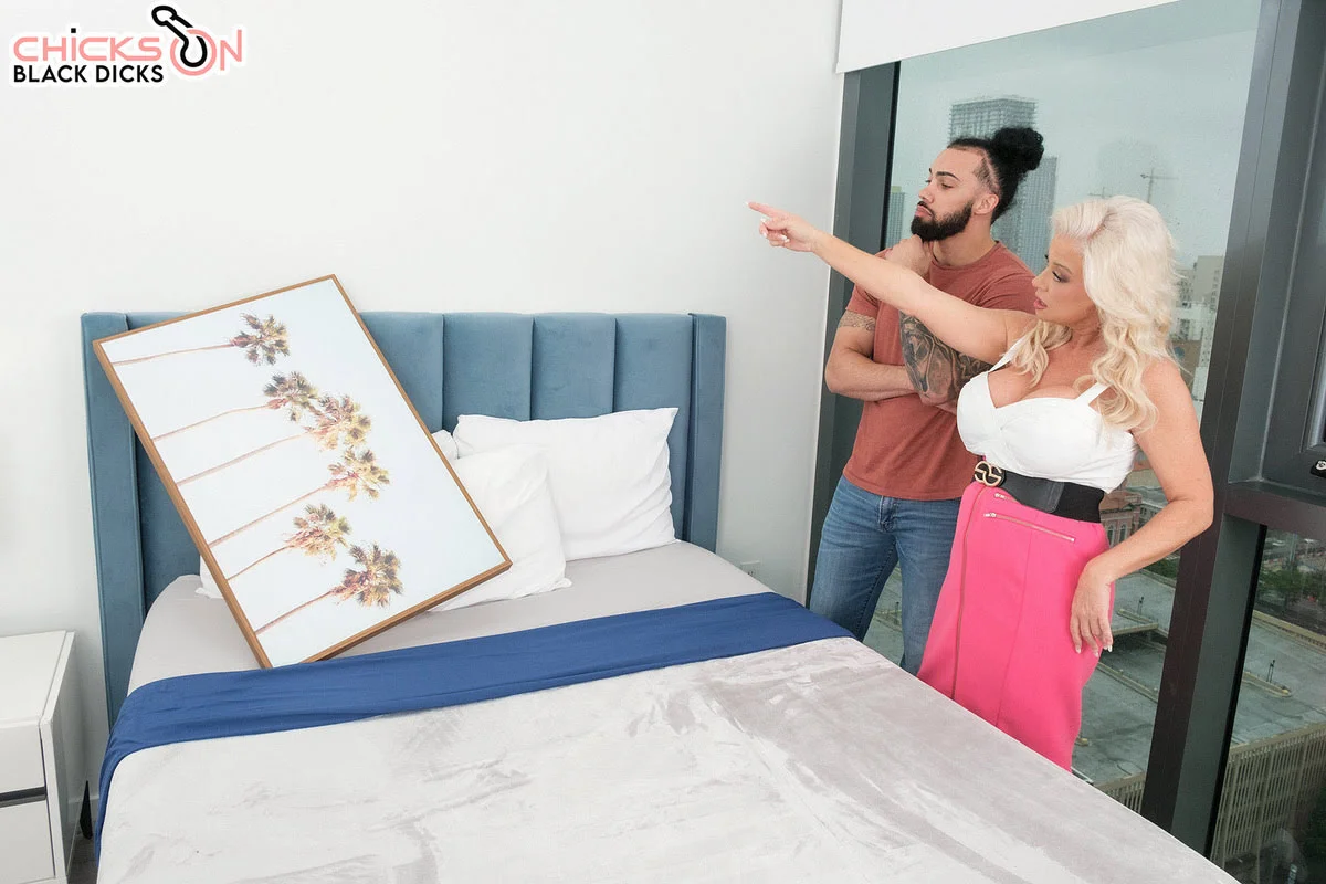 Busty blonde MILF Billie Jean Austin in white top and pink skirt points dramatically in modern bedroom with James Angel, tattooed man in man bun, close behind her